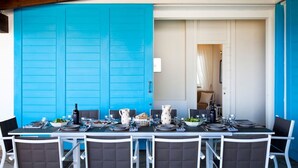 Un coin repas avec une longue table dressée pour six personnes, des chaises blanches et, au fond, une porte d’un bleu vif.