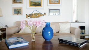 Un salon avec un canapé beige, une table basse sur laquelle sont posés des livres et un vase de fleurs, et des tableaux de paysages encadrés accrochés au mur.
