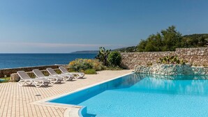 Un espace piscine doté de chaises longues et d’un mur en pierre, avec vue sur la mer.