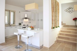A modern kitchen with white cabinetry, a central island with bar stools, a built-in oven, and a stainless steel range hood. - Villa Bianca - Four Bedroom Villa, Sleeps 12 (Melissano)