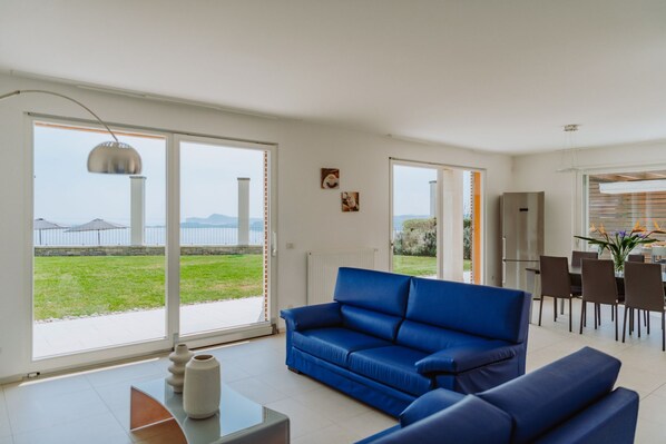A modern living room with a blue sofa, a dining area with chairs, and a kitchen with a refrigerator. - Casa Delle Arche - Five Bedroom Villa, Sleeps 10 (Gardone Riviera)