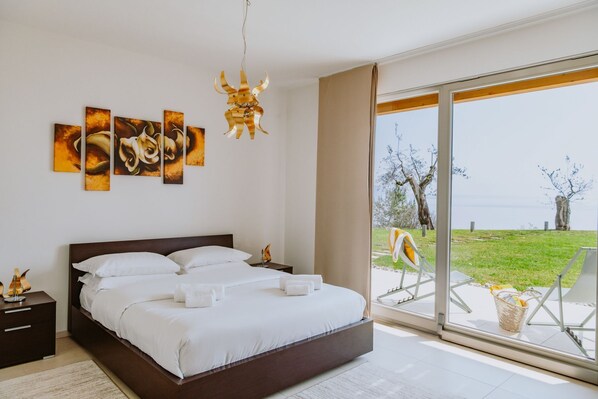 A bedroom with a large bed, a nightstand, a wall-mounted lamp, and a view of the outdoors through a sliding glass door. - Casa Delle Arche - Five Bedroom Villa, Sleeps 10 (Gardone Riviera)