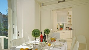 Mesa de comedor servida con platos, vasos y una cesta de frutas, con vista a un balcón y una cocina al fondo.