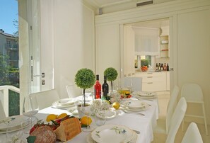 A dining table set with plates, glasses, and a fruit basket, with a view of a balcony and a kitchen in the background. - Bianca Neve - Five Bedroom Villa, Sleeps 11 (Toscolano Maderno)