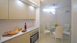 Una cocina moderna con armarios blancos, una mesa de comedor de vidrio y sillas blancas. Hay una barra de pan, una botella y una tabla de cortar sobre la encimera.