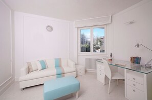 A modern, white-themed room with a glass desk, a white chair, a white sofa, and a small bench.