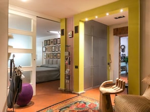 Una stanza con un letto, un armadio, un tavolino e una sedia. Si apre una porta verso un'altra stanza con un letto e una parete decorata con quadri.