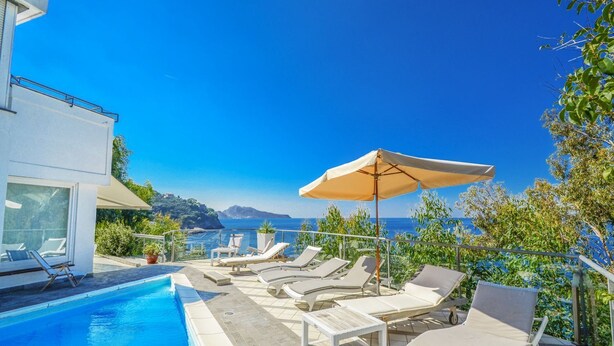 A poolside area with lounge chairs and umbrellas, overlooking a clear blue sea and distant mountains.