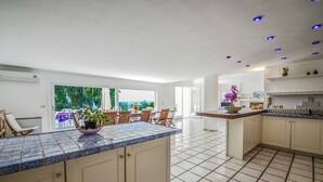 A spacious kitchen with a tiled floor, a central island with a bowl of flowers, and a dining area with chairs and a table.