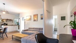 A modern living room with a grey sofa, a black armchair, and a wooden coffee table. There is a kitchen area with white cabinets and a microwave visible in the background. - House heated pool walking beach animals Ok (Labenne)