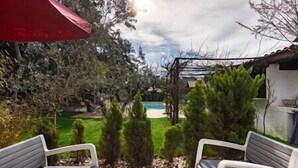 A patio with two white chairs, a small table, and a red umbrella. In the background, there is a swimming pool surrounded by greenery and a building. - House heated pool walking beach animals Ok (Labenne)