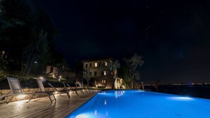 A nighttime swimming pool with a wooden deck and lounge chairs, surrounded by trees and a building with illuminated windows.