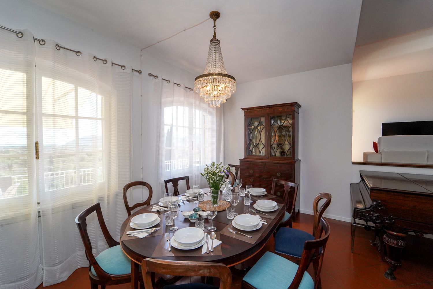 A dining room with a round table set for six, surrounded by wooden chairs with blue cushions. A chandelier hangs from the ceiling, and a wooden cabinet with glass doors is visible in the background.