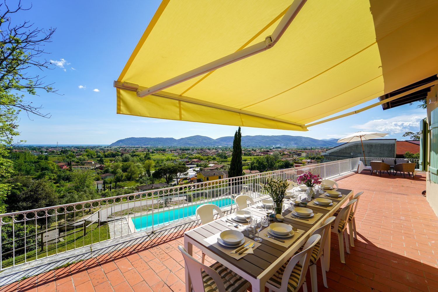 A terrace with a dining table set for four, overlooking a swimming pool and a scenic landscape.