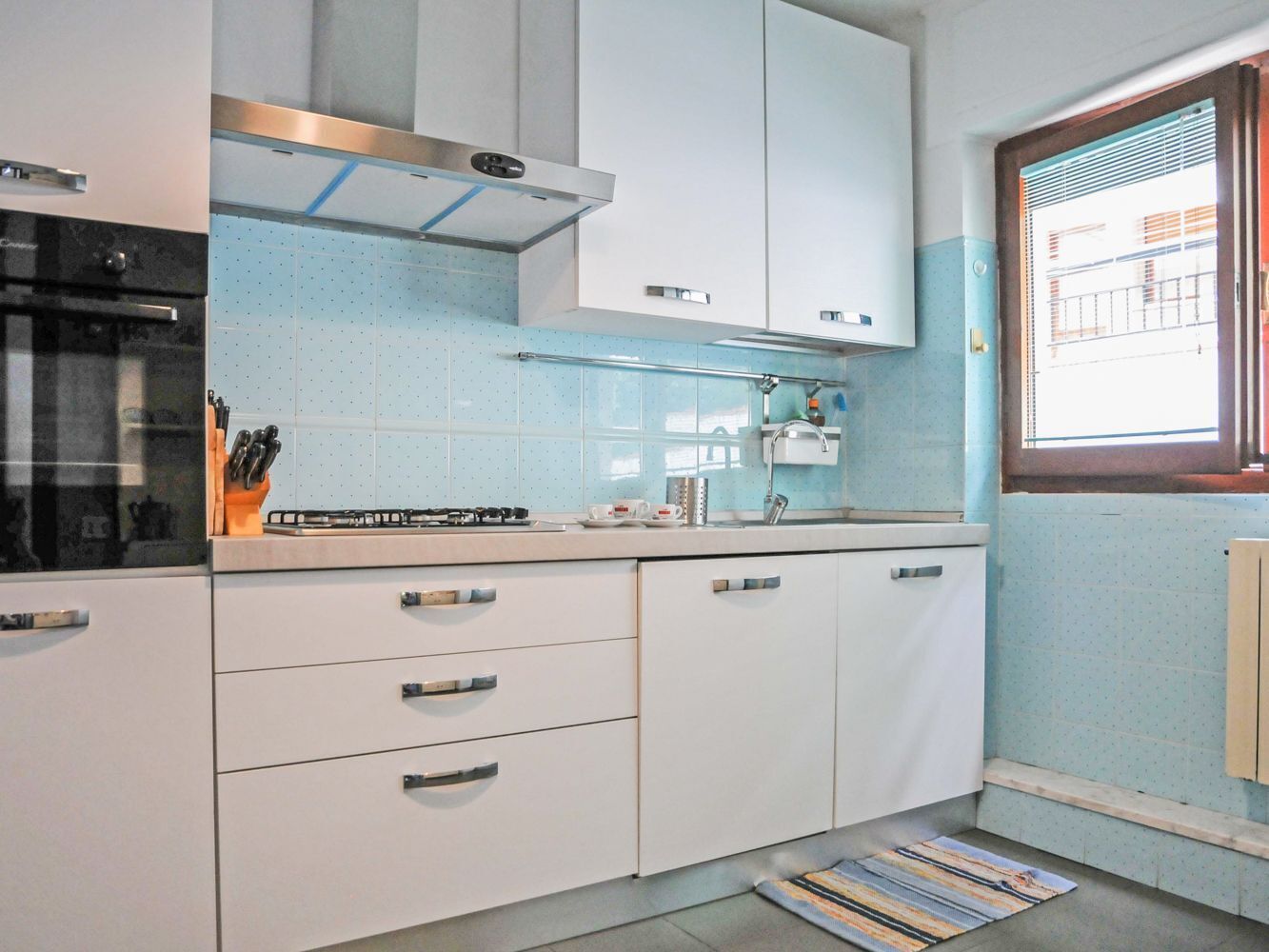 Une cuisine avec des meubles blancs, une cuisinière et une fenêtre avec des stores.