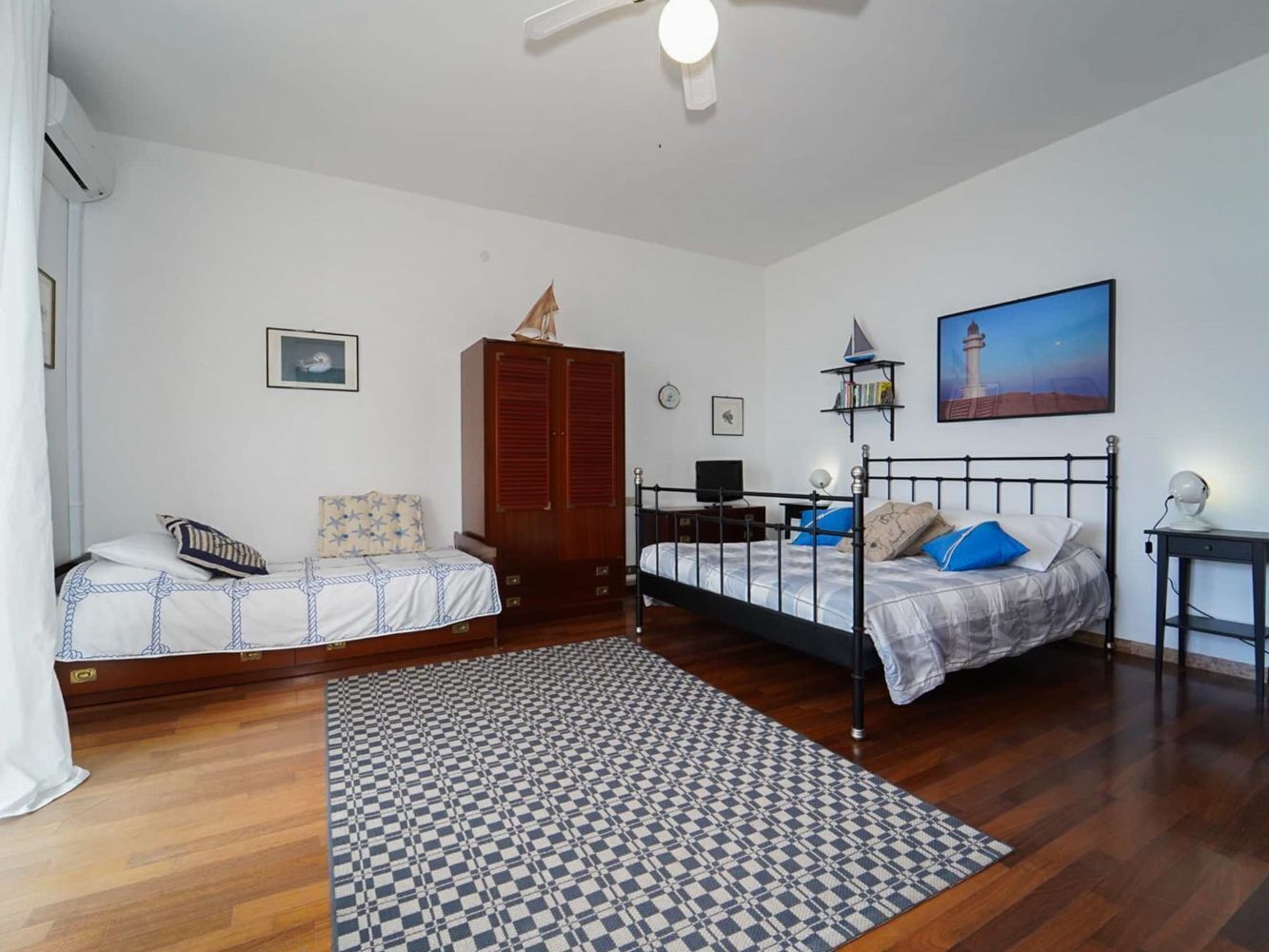 A bedroom with two beds, a wooden wardrobe, a desk, a chair, a lamp, and a painting on the wall.