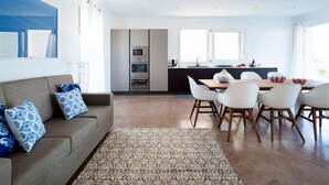Un salon moderne comprenant un canapé, une table à manger avec des chaises, un coin cuisine équipé d’appareils électroménagers et une grande fenêtre laissant entrer la lumière naturelle.