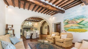 Un soggiorno spazioso con soffitto alto, travi in legno, zona pranzo e un affresco raffigurante un paesaggio.