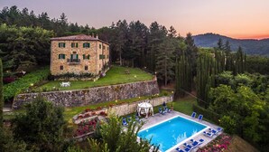Un edificio in pietra con piscina e lettini, circondato da vegetazione lussureggiante e vista sulle colline.