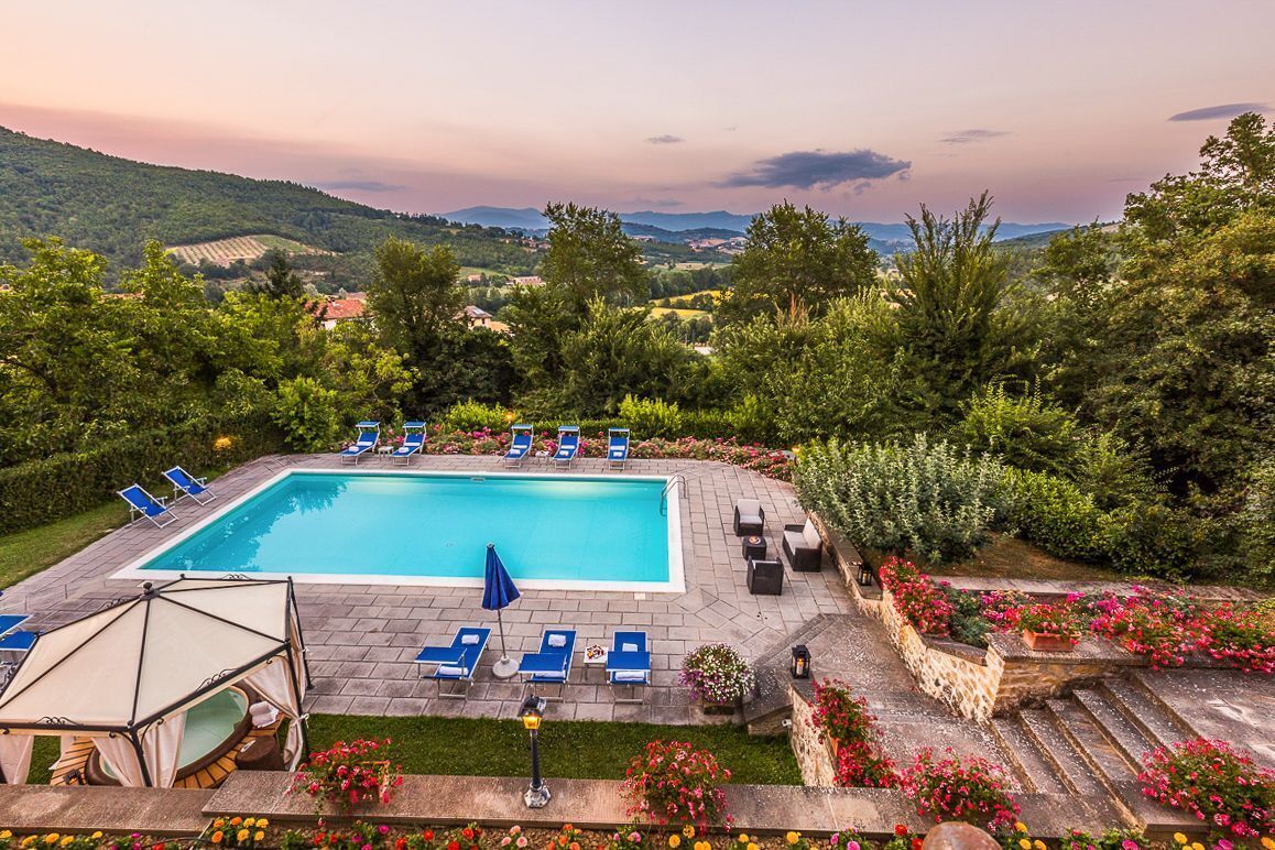 A rectangular swimming pool surrounded by lounge chairs and a covered seating area, with a view of a hilly landscape in the background.