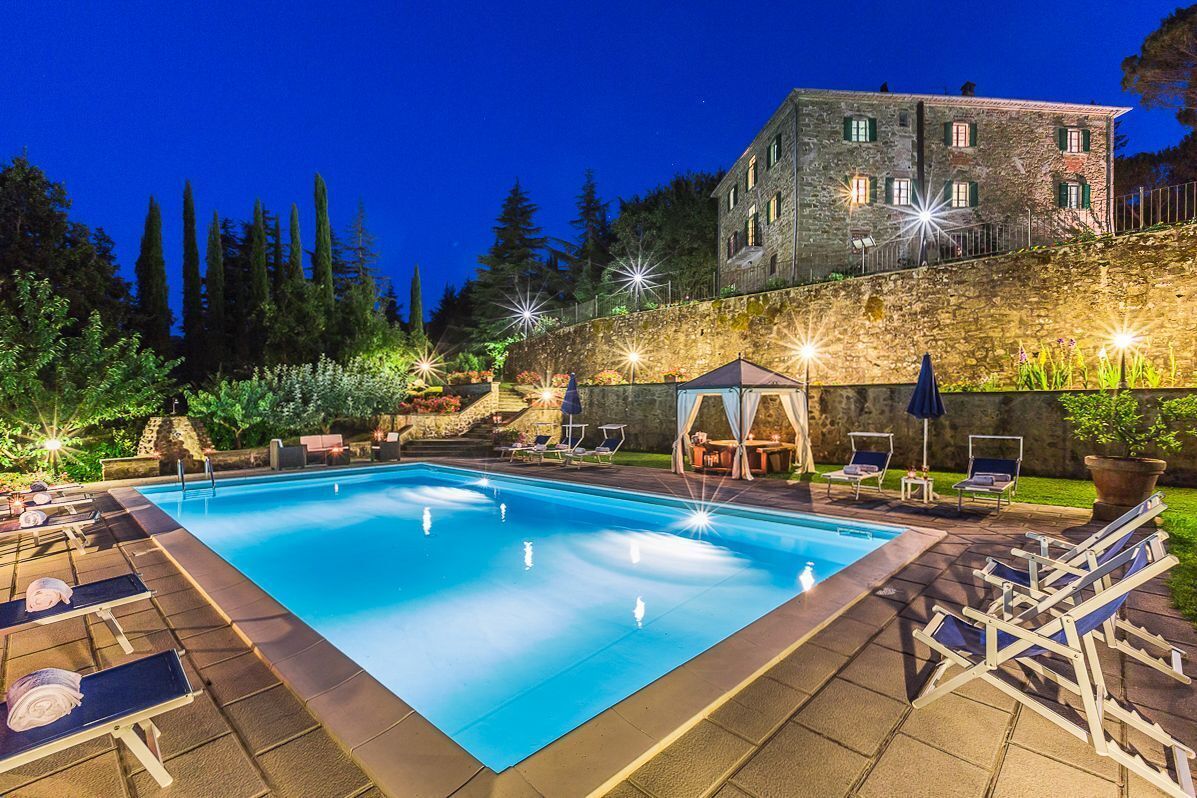 A rectangular swimming pool with lounge chairs and umbrellas, surrounded by a stone wall and a building with multiple windows.