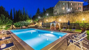 Una piscina rettangolare con lettini e ombrelloni, circondata da un muro in pietra e un edificio con diverse finestre.