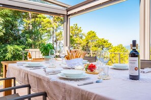 Un espace de restauration en plein air avec une table dressée pour deux personnes, agrémentée d’une bouteille de vin, de verres, de pain et d’un saladier de fruits.