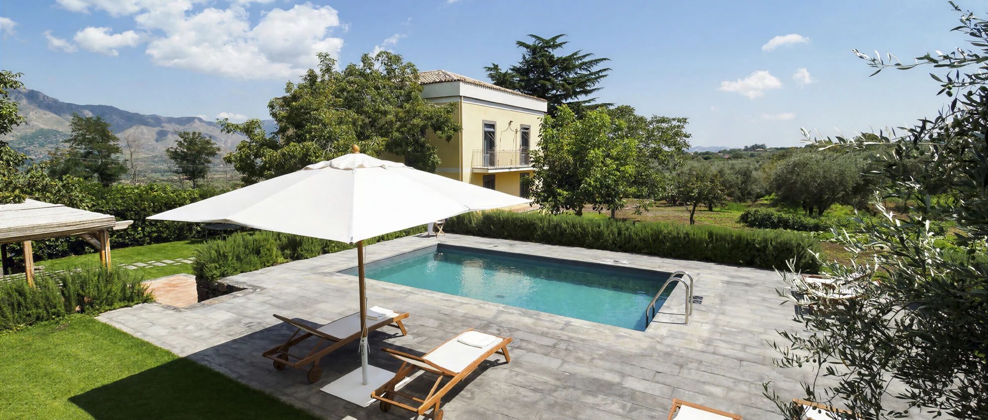 Una piscina con pérgola y sillas de descanso, rodeada de un jardín ajardinado y un paisaje montañoso.