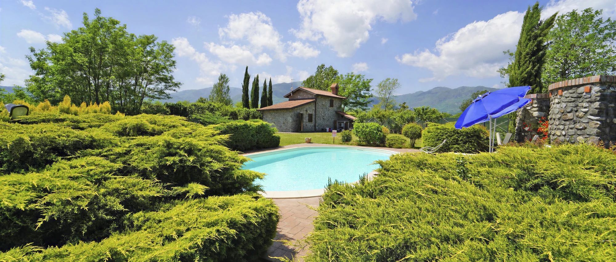 A swimming pool surrounded by lush greenery, a stone building, and a mountain backdrop.