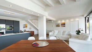 A modern kitchen with a central island, a dining area with a wooden table, and a living room with a white sofa and a large window. - Cella Mimavi - Three Bedroom Villa, Sleeps 6 (Ribera)