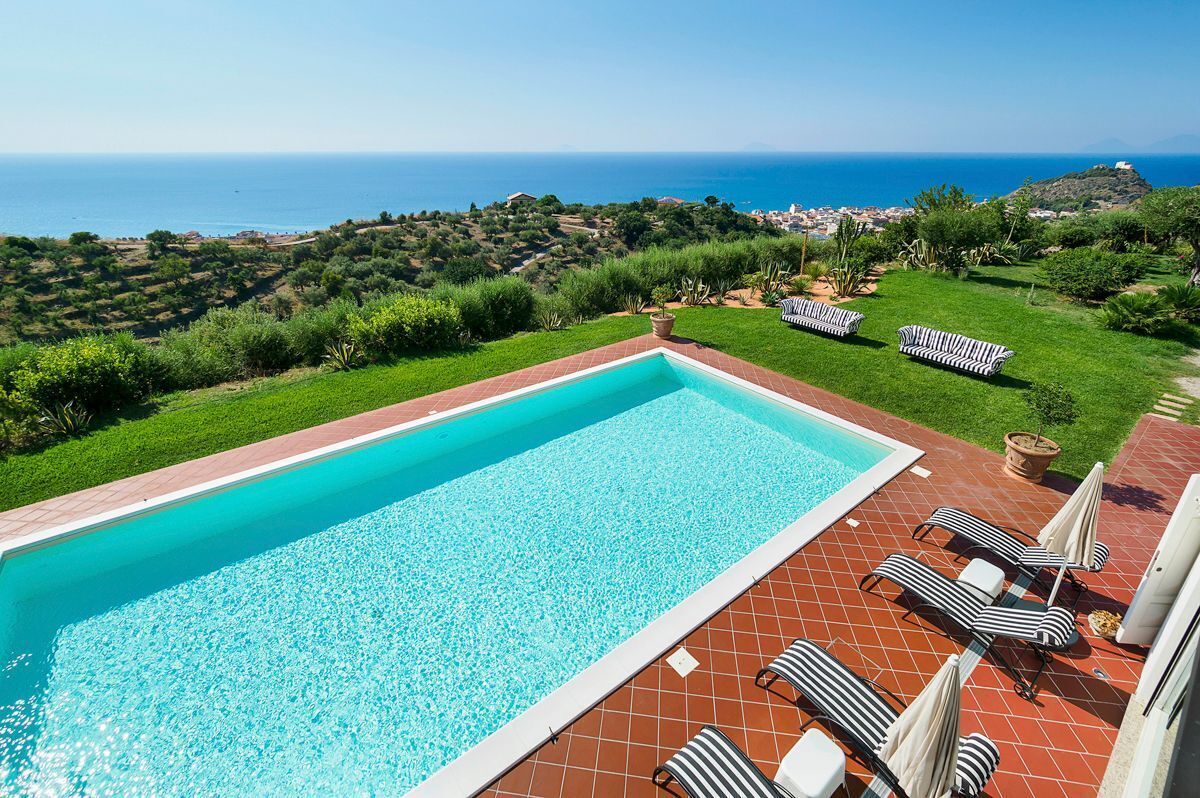 A rectangular swimming pool with a clear blue water, surrounded by a red-tiled deck. There are striped lounge chairs and a small potted plant on the deck.