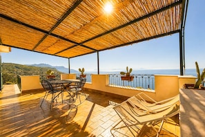 Eine überdachte Terrasse im Freien mit Tisch und Stühlen, Topfpflanzen und Blick auf das Meer und die Berge.