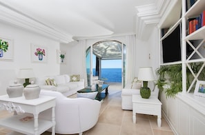 A spacious living room with a white color scheme, a large window offering a view of the sea, and a bookshelf filled with books.