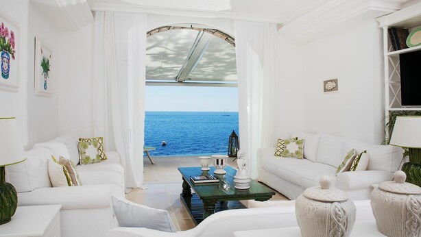 Un salón blanco con vistas al mar, que incluye un sofá, una mesa de centro y cojines decorativos.