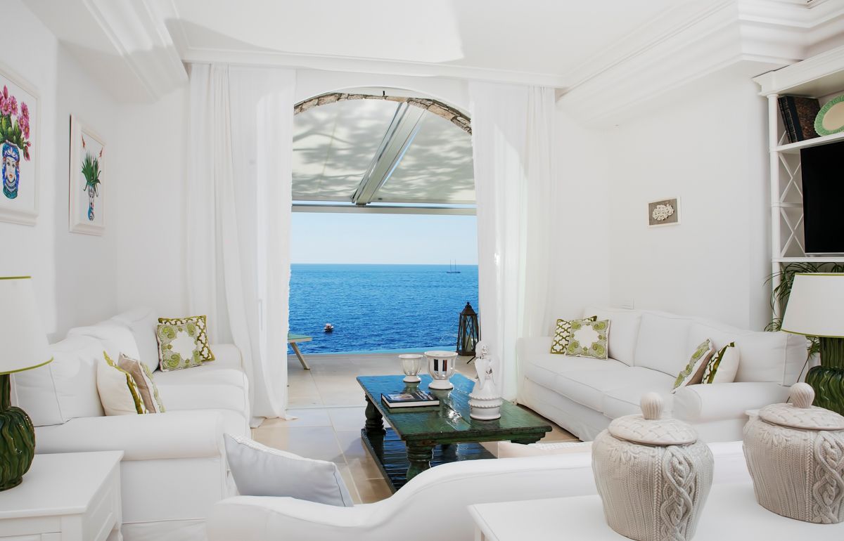 A white living room with a view of the sea, featuring a sofa, a coffee table, and decorative pillows.