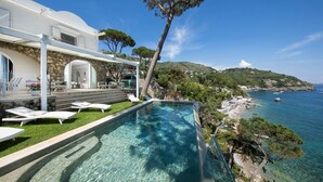 Zona junto a la piscina con sillones de descanso, un muro de piedra y vistas al mar y colinas.