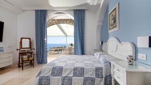 Un dormitorio con una cama grande, un aparador, un espejo y vista al mar a través de un ventanal de arco.