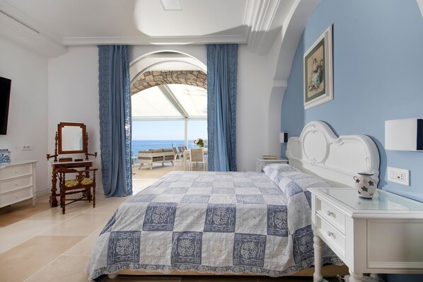 Une chambre à coucher avec un grand lit, une commode, un miroir et une vue sur la mer, visible à travers une fenêtre en arc de cercle.