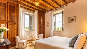 Un dormitorio con una cama grande, vigas de madera en el techo, una mesita con una silla y una ventana con vista a árboles.