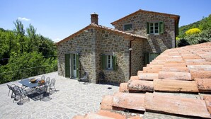A stone house with a tiled roof, a patio with a table and chairs, and a view of the surrounding greenery.