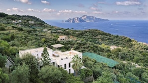 Una casa moderna con techo plano y un balcón amplio, rodeada de vegetación exuberante y con vistas a la costa y montañas distantes.