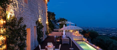 Una terraza con piscina, mobiliario de exterior e iluminación con luces colgantes.