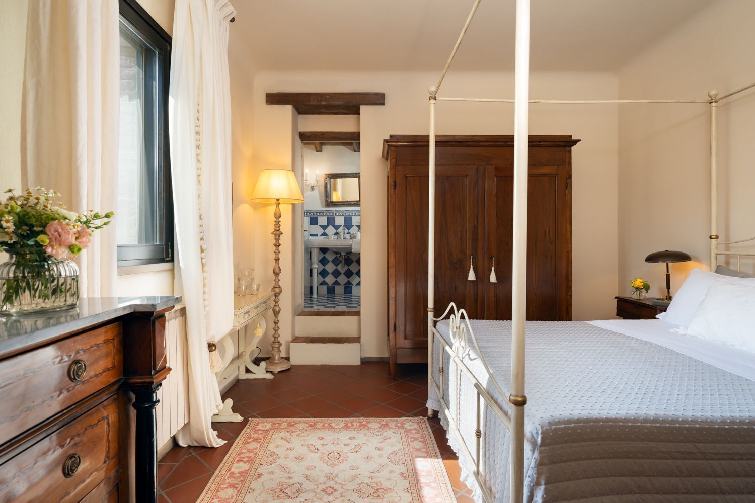 A bedroom with a four-poster bed, a wooden dresser, a nightstand with a lamp, and a bathroom with a blue and white checkered tile backsplash.