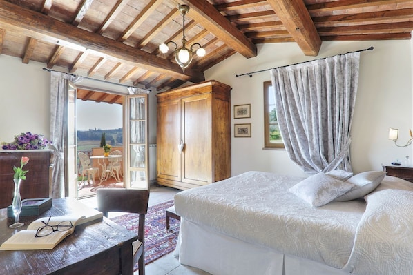 Une chambre avec un grand lit, des poutres apparentes en bois au plafond, une armoire et une vue sur l’extérieur.