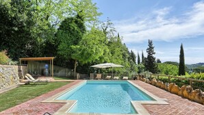 Una piscina rectangular con un borde empedrado, rodeada por un muro de piedra y abundante vegetación. Hay sillas de descanso y una mesa con una bebida y una flor en el borde.