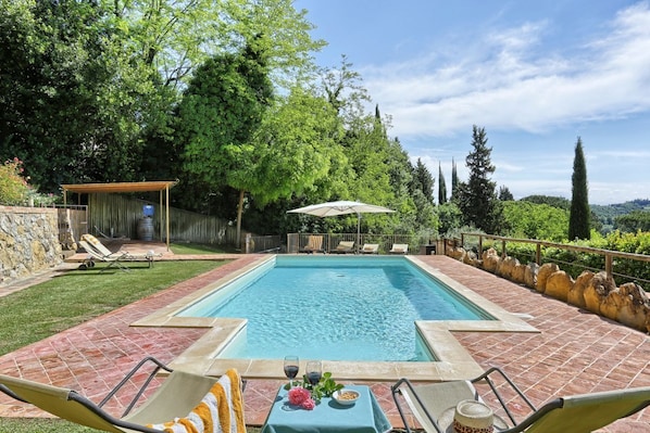 Une piscine rectangulaire, bordée d’une terrasse carrelée, entourée d’un mur de pierre et d’une végétation luxuriante. On y trouve des chaises longues et une table sur laquelle sont posés une boisson et une fleur.