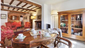 Un área de comedor con una mesa de madera dispuesta para el té, rodeada de sillas tapizadas en rojo. Se observan un hogar a leña de ladrillo y un mueble de madera con puertas de cristal al fondo.