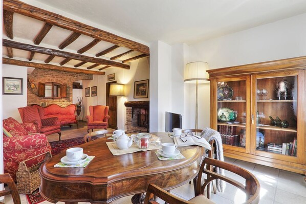 Un coin repas avec une table en bois mise pour le thé, entourée de chaises à revêtement rouge. On aperçoit au fond une cheminée en briques et un buffet en bois doté de portes en verre.