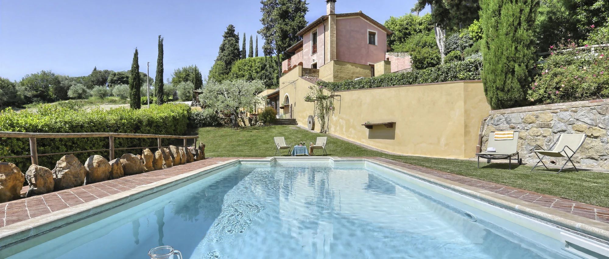 A swimming pool with a stone wall and lounge chairs, surrounded by greenery and a building with a red roof.