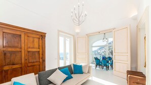 A modern living room with a beige sofa, blue cushions, a wooden cabinet, and a view of the city through an open door. - Villa Del Piano - Four Bedroom Villa, Sleeps 8 (Amalfi)